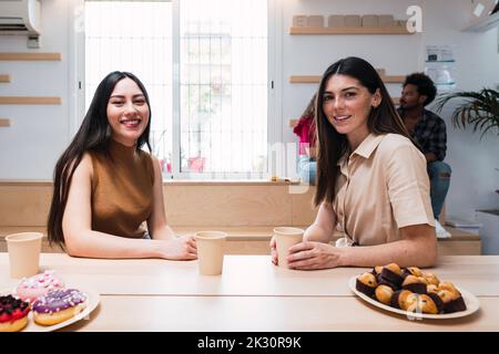 Smiling young businesswomen with disposable coffee cups sitting at table in cafeteria Stock Photo