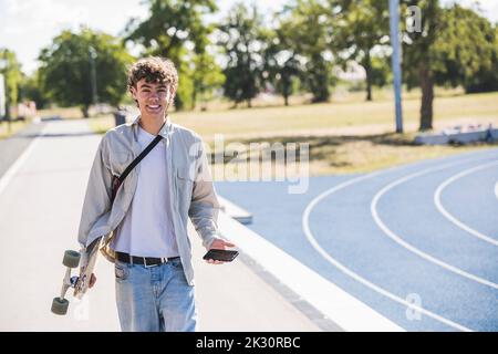 Happy young man holding skateboard and mobile phone on footpath Stock Photo