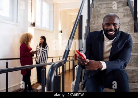 Smiling businessman with smartphone sitting on stairs colleagues talking in background Stock Photo