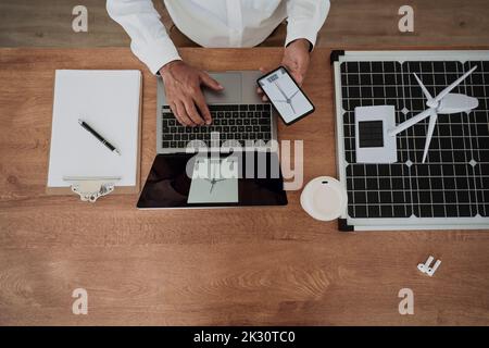 Hands of architect holding smart phone using laptop sitting by wind turbine on solar panel at desk Stock Photo