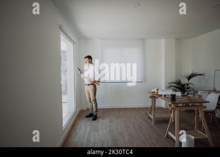 Architect holding rolled up blueprint using smart phone standing by window in office Stock Photo