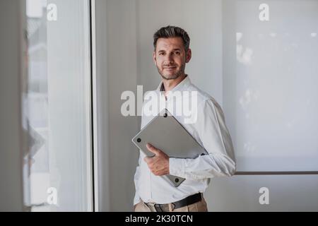Smiling businessman holding laptop standing by window in office Stock Photo