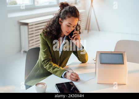 Businesswoman talking on smart phone writing at desk in office Stock Photo