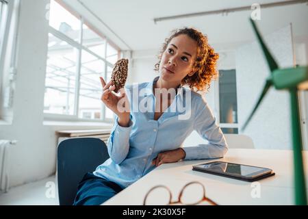 Thoughtful engineer with cookie sitting by wind turbine model on desk Stock Photo