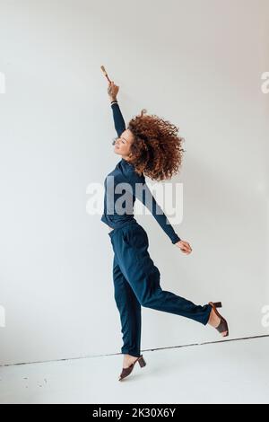 Cheerful young woman with hand raised jumping in front of white wall Stock Photo