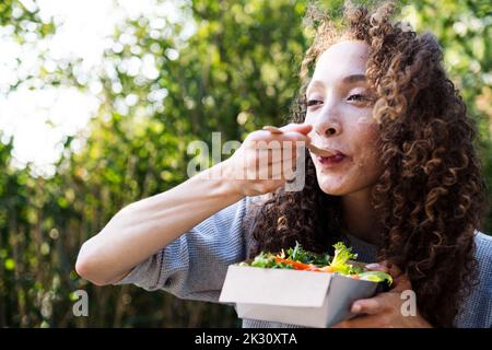 Young woman eating vegetable salad in park on sunny day Stock Photo