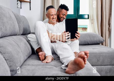 Mature man using tablet PC sitting with boyfriend on sofa at home Stock Photo