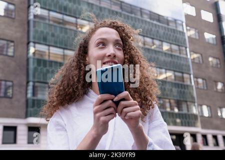 Contemplative woman with curly hair holding smart phone in front of building Stock Photo