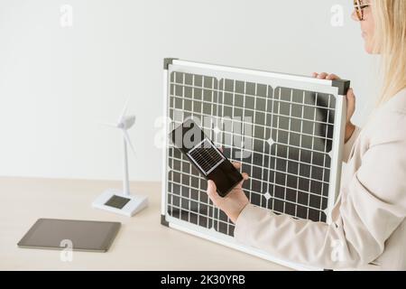 Woman holding mobile phone in office with solar panel and wind turbine model on desk Stock Photo