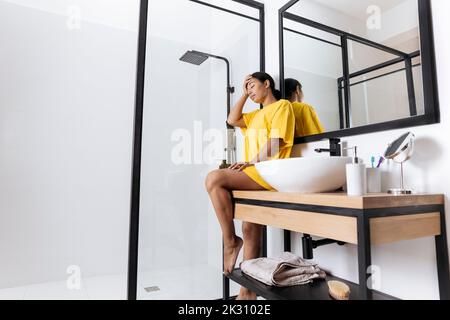 Upset woman touching holding head in hand sitting by bathroom sink Stock Photo