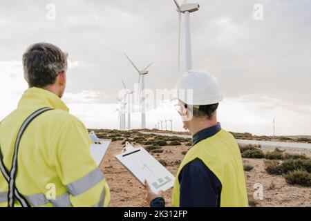 Technicians holding clipboards looking at wind turbines at wind farm Stock Photo
