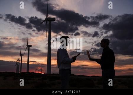 Engineers discussing with each other holding laptop at wind farm on sunset Stock Photo
