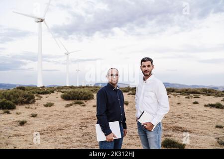 Confident engineers with tablet PC and laptop standing at wind farm Stock Photo