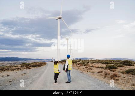 Engineers standing on dirt road looking at wind turbines at wind farm Stock Photo