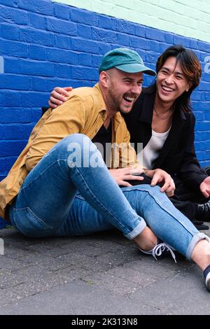 Happy gay couple sitting on sidewalk in front of wall Stock Photo