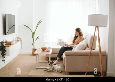Young woman with dog using laptop on sofa at home Stock Photo