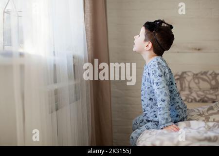 Boy with sleep mask looking through window sitting on bed Stock Photo