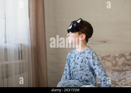 Cute boy with sleep mask looking through window sitting on bed Stock Photo