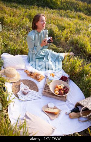 Laughing woman in a long dress with short hair sitting on a white blanket with fruits and pastries and looking at the pictures on camera. Concept of h Stock Photo