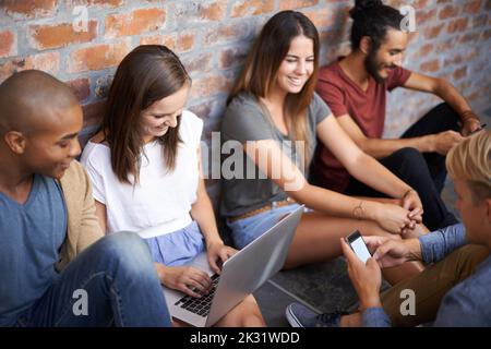 Its fun working in a laid-back environment. A diverse group of friends sitting in a hallway and using their digital devices. Stock Photo