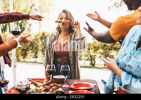 Friends having fun party pointing a curvy young woman making faces holding skewers grilled on barbecue - people gathering at weekend eating together a Stock Photo