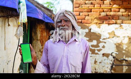 Indian old man holding a mobile phone and point towards it's screen. Mobile phone with a green screen for mockup. Stock Photo