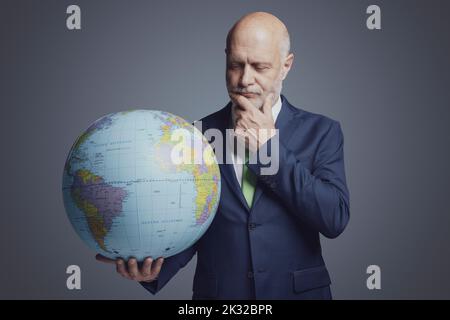 Corporate business executive holding a globe and thinking with hand on chin, global business responsibility and solutions concept Stock Photo