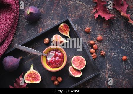 Autumntime background with fig jam, spoon, nuts and fresh halved fig fruits on black wooden tray. Dark brown background with dry Fall leaves. Stock Photo