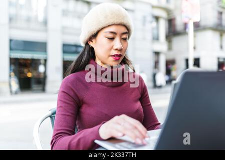 Pensive Chinese female browsing internet on modern netbook while sitting at table on terrace of cafe on street in city Stock Photo