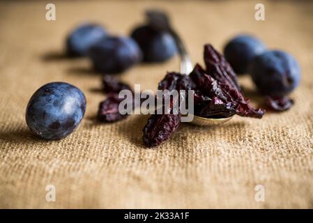 Dried prunes in a spoon on a burlap tablecloth against a background of fresh plums. Prunes and plums on a kitchen table close-up. Stock Photo