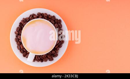 Cup of coffee with pink foam on a coral background. Coffee cup and roasted coffee beans on a saucer. Top view. Copy space Stock Photo