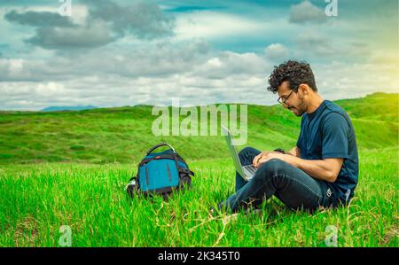 Smiling freelancer young man with laptop working from the field. Close up of man sitting on green grass working with his laptop. Smiling man with Stock Photo