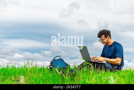 Smiling man with laptop sitting on the grass in the field. Side view of man sitting on green grass working with his laptop. Smiling freelancer guy Stock Photo