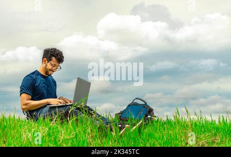 Smiling freelancer man sitting with laptop and backpack in field. Portrait of a man sitting on the grass of the field working with his laptop. Stock Photo