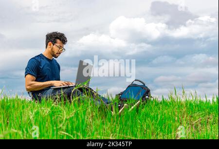 Freelancer man sitting with laptop and backpack in the field. Man sitting in the green field working from his laptop. Freelancer man concept working Stock Photo