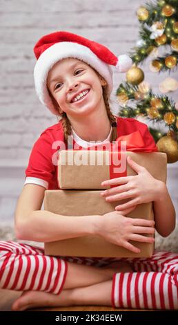 Portrait of a Christmas happy smiling girl in a red hat, holding a Christmas gift in her hands and looking into the camera. Sitting in a room on the Stock Photo