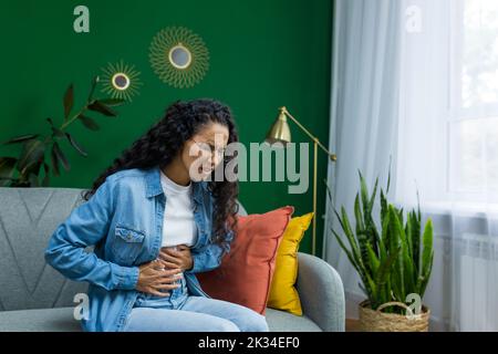 Woman sick alone at home, Hispanic woman sitting on sofa in living room having severe stomach pain. Stock Photo