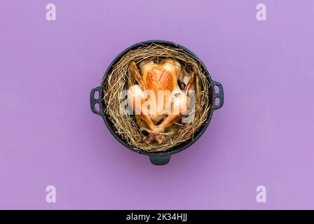 Above view with a whole chicken baked in a rustic method, on a layer of hay in an iron cast pot. Oven baked chicken, top view minimalist on a purple b Stock Photo