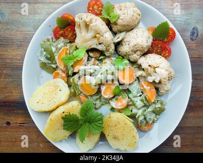 Top view of a platter of steamed fresh vegetables, cauliflower, sliced carrots, and chopped green beans rolled in a light herb cream. Sliced jacket po Stock Photo