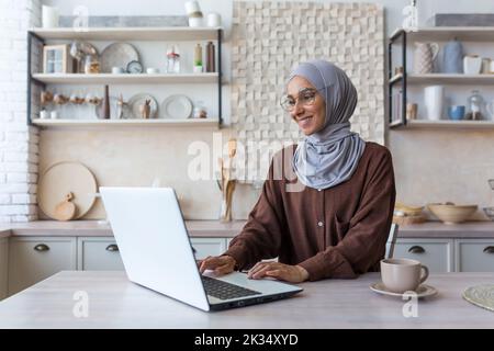Young beautiful Muslim woman in hijab working remotely from home, using laptop while sitting in kitchen at table, woman in glasses smiling and happy with work. Stock Photo