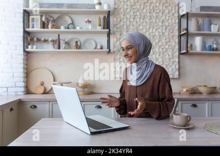 Young beautiful Muslim woman in hijab using laptop for video call, woman at home sitting in kitchen talking with friends remotely smiling at computer web camera Stock Photo