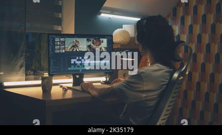 Male editor in headphones sitting at the table at night and editing video with astronauts for customer on pc in professional program while working remotely Stock Photo