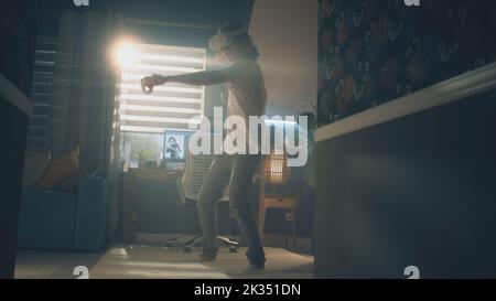 Guy moving while playing video game at home using VR headset and wireless controllers in spare time. Virtual reality device. Cyberspace and metaverse Stock Photo
