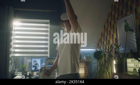 Guy moving while playing video game at home using VR headset and wireless controllers in spare time. Virtual reality device. Cyberspace and metaverse Stock Photo