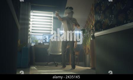 Guy moving while playing video game at home using VR headset and wireless controllers in spare time. Virtual reality device. Cyberspace and metaverse Stock Photo