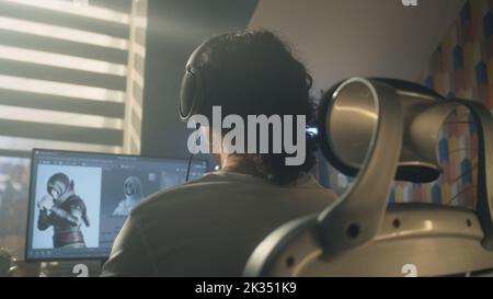 Back view of man in headphones sitting at the table and creating video game hero and 3D animation in professional computer program, while working remotely at home. Stock Photo