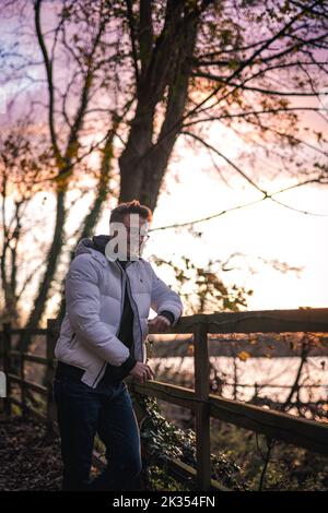 A vertical of a Caucasian man in glasses leaning on a wooden fence in a park captured at sunset Stock Photo