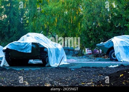lightly armored military equipment covered with tarpaulin stands in the city park during repair work, selective focus Stock Photo