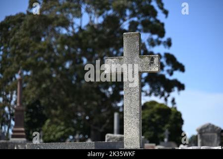 Weathered stone cross in a cemetery on a sunny day, with graves, a large tree, and blue sky in the background Stock Photo