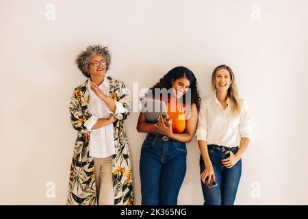 Successful businesswomen laughing happily while standing against a wall background. Group of three multicultural businesswomen working together in an Stock Photo